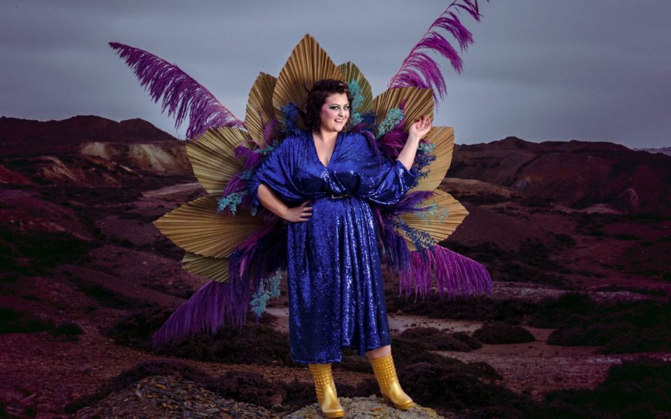 Kiri Pritchard-Mclean standing on a rock in a moody outdoor landscape. She wears a blue dress and gold wellies with large purple and gold feathers all around her.