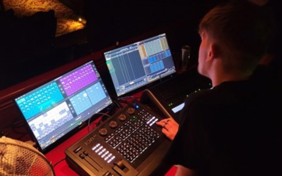 A over the shoulder shot of a student sat at the lighting desk turning dials and pressing buttons in front of two screens.