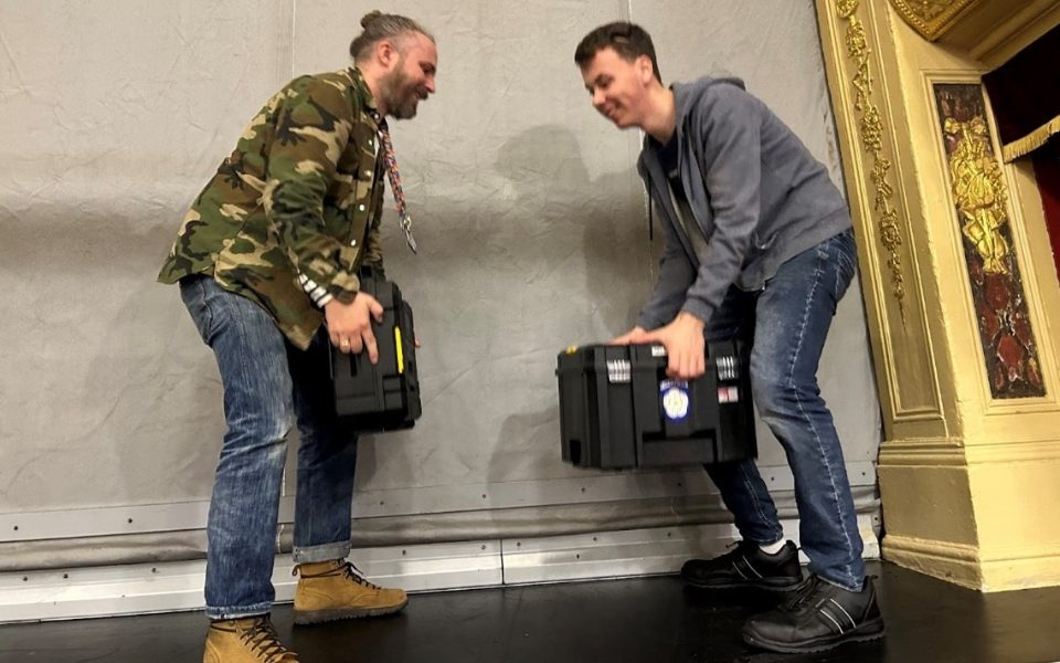 Two men lifting tool boxes facing each other with their knees bent.