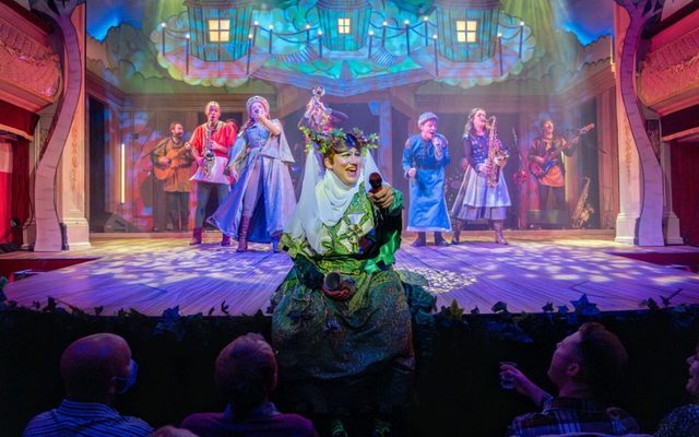 Simon Nock (Nanny Nellie) and cast Simon Nock (Nanny Nellie) sits on stage addressing the audience, cast behind him. Credit Ant Robling