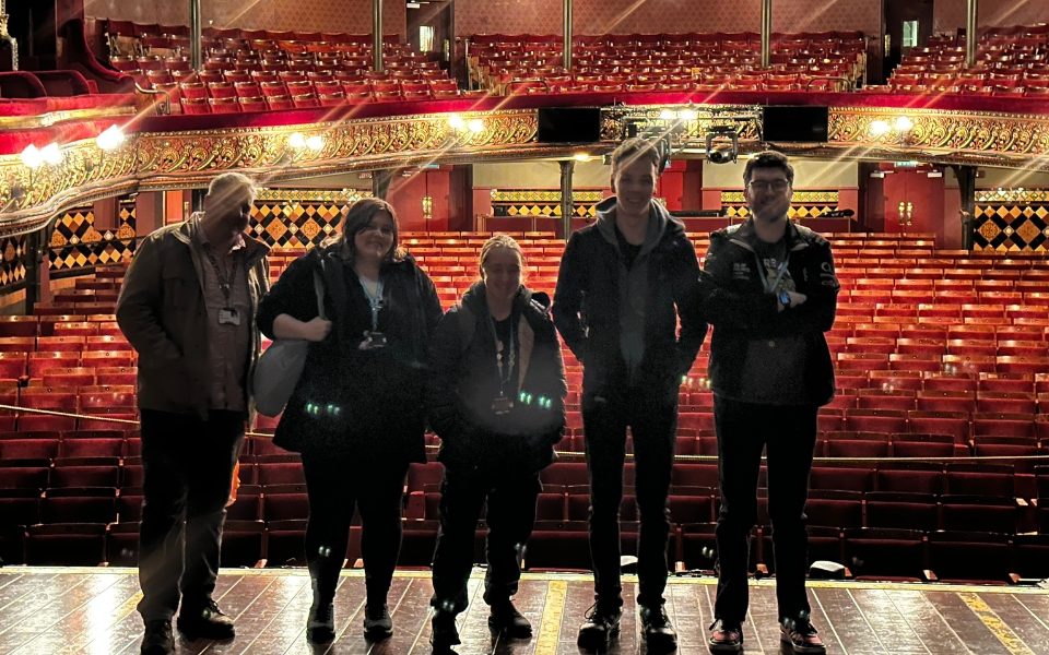 Five people standing on the Leeds Grand Theatre stage smiling at the camera. Behind them is the auditorium with rows of red chairs.