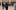 A group of people standing in a circle. They are standing with a finger on their chin and looking up mischievously.