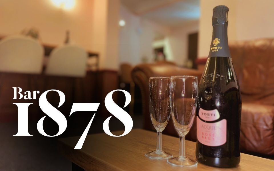 A bottle of pink Prosecco and two champagne flutes stand in front of a blurred background of a bar.