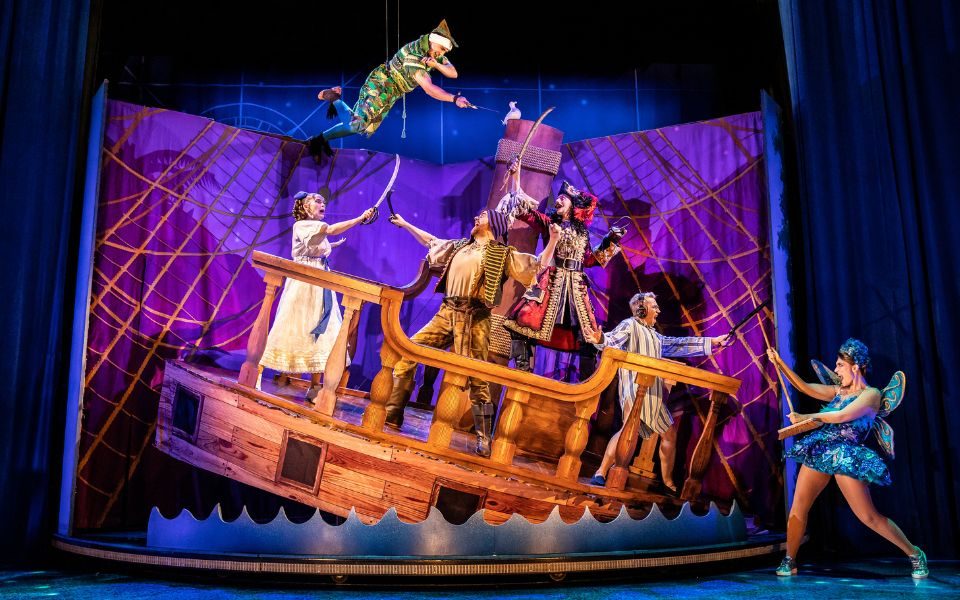 A scene from Peter Pan Goes Wrong with a boat set piece at an angle going into the water. The characters are swordfighting while Peter Pan flies on a wire over them.