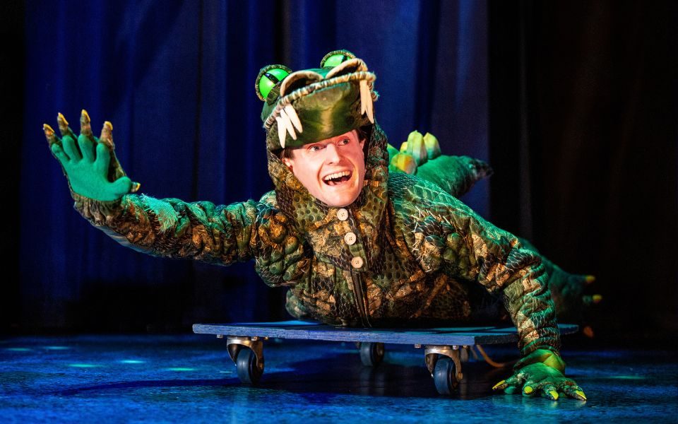An actor dressed as a crocodile lying on a skateboard and waving