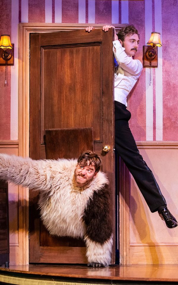 A production photo from Peter Pan Goes Wrong. A man hangs off the top of a wooden door while a man dressed as a dog crawls through a hole in the bottom of it.