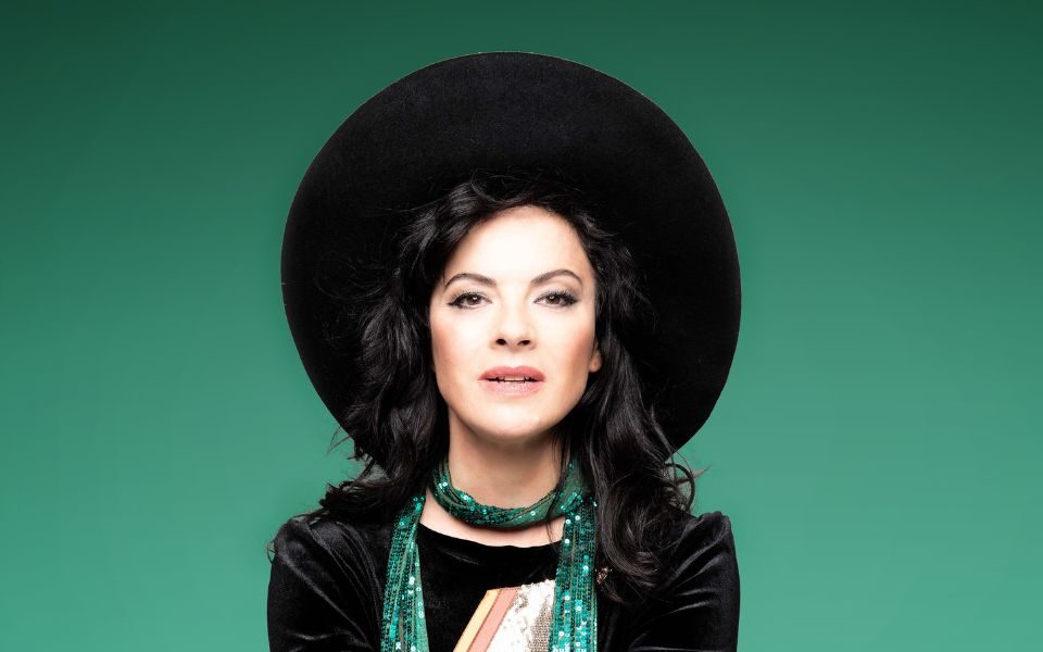 Camille O'Sullivan wearing a big black hat and green sparkly scarf sat in front of a green background.