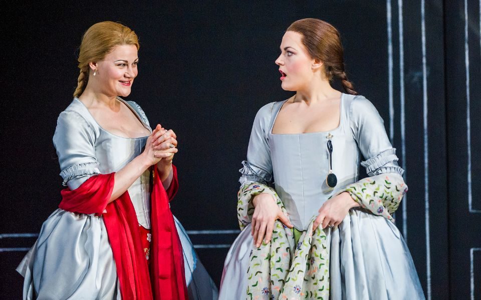 Helen Sherman as Dorabella and Máire Flavin as Fiordiligi look at each other wearing grey 18th century dresses. Dorabella has a bright red scarf over her arms.