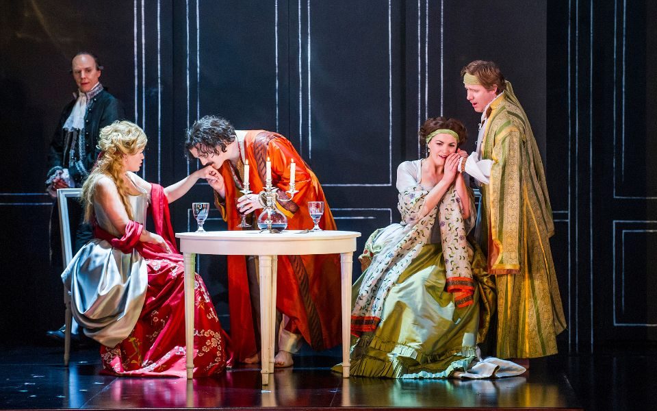 Don Alfonso watches in the background as the lovers are onstage in brightly coloured 18th century clothing. The women sit at a table with glasses and candles. Guglielmo kisses Dorabella's hand and Fiordiligi cradgles Ferrando's hand to her face.