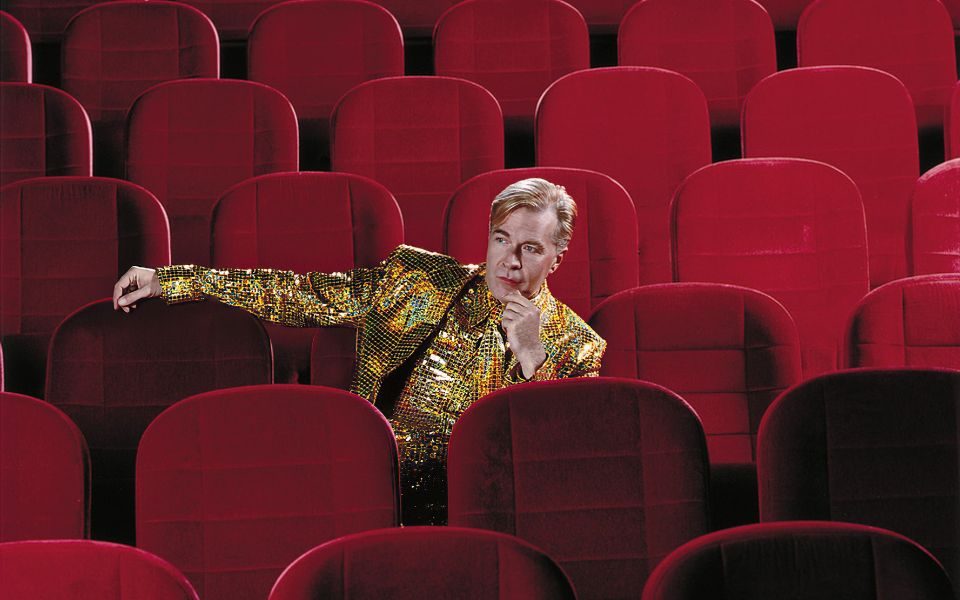 Martin Fry sat in a middle row of some traditional red theatre or cinema auditorium seats.