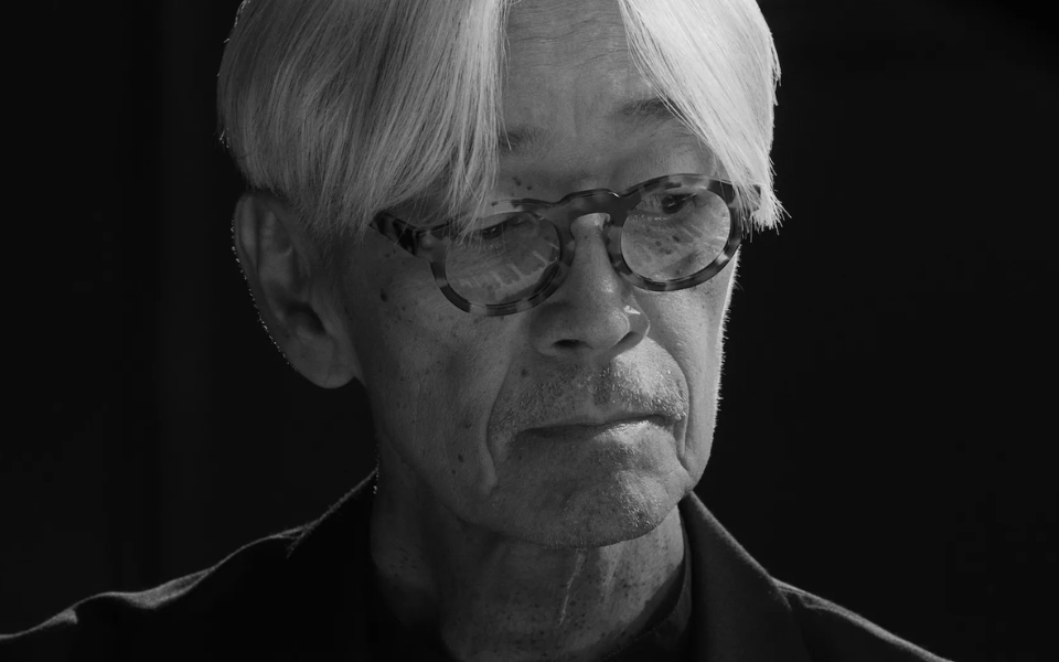 Black and white image of Ryuichi Sakamoto from the shoulders up wearing glasses.