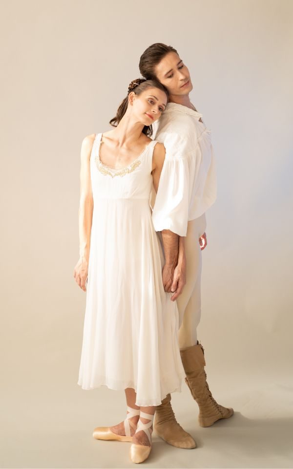 Amber Lewis and Joseph Taylor standing back to back and leaning their heads on each other. They stand in a studio in their white costumes for Romeo and Juliet.