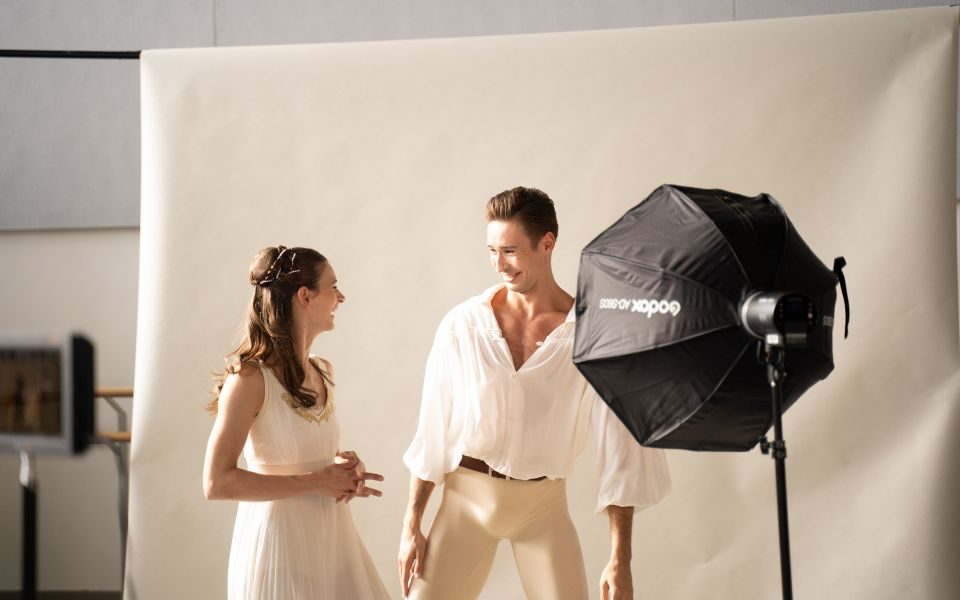 Amber Lewis and Joseph Taylor looking at each other and laughing. They wear light coloured costumes in front of a cream backdrop at the Romeo & Juliet photoshoot.