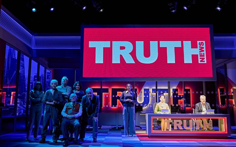 The cast of Drop The Dead Donkey on stage with a giant red screen reading Truth News above a news desk.