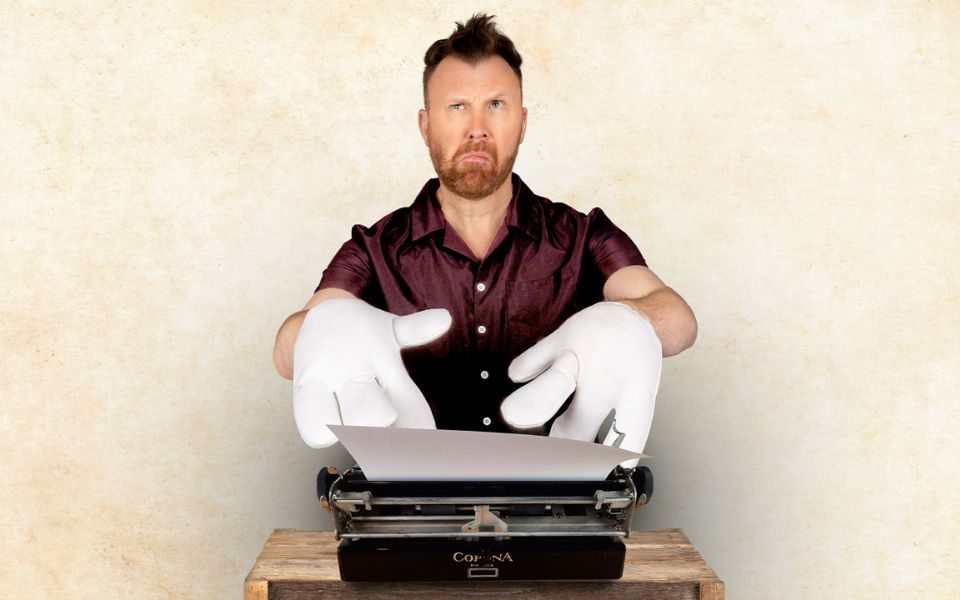 Jason Byrne wearing chunky white gloves typing on a typewriter looking confused.