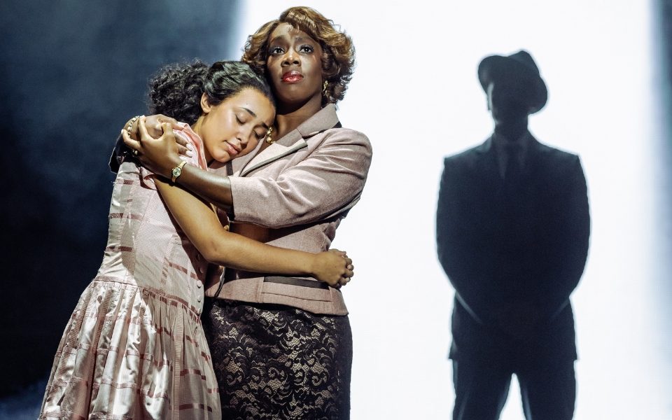 Jaydah Bell-Ricketts as Girl and Carly Mercedes Dyer as Faye Treadwell hugging on stage with Miles Anthony Daley's silhouette as George Treadwell appearing in the background.