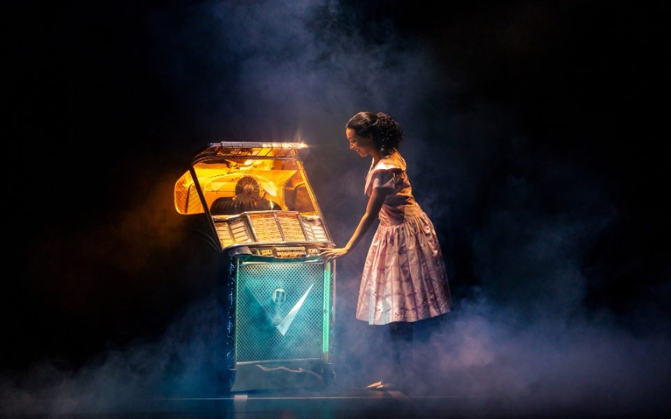 Jaydah Bell-Ricketts as Girl on stage in The Drifters Girl stood at a lit up old fashioned jukebox.