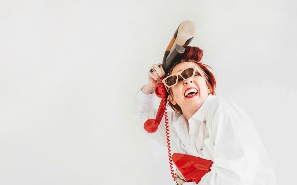 Laura Belbin in a white shirt holding a red cord telephone to her ear with the same hand as holding a high heel, the dialler is in her other hand and she has sunglasses falling off her eyes.