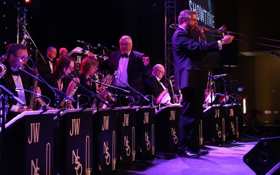 Members of the Northern Swing Orchestra on stage including the conductor and a trombonist standing up to do a solo.