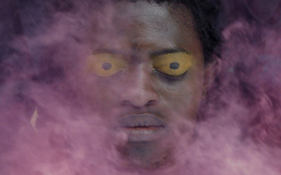 Image of a person's head with closed eyes and yellow eyes painted on their eyelids. Their face is obscured with pinky-purple smoke.