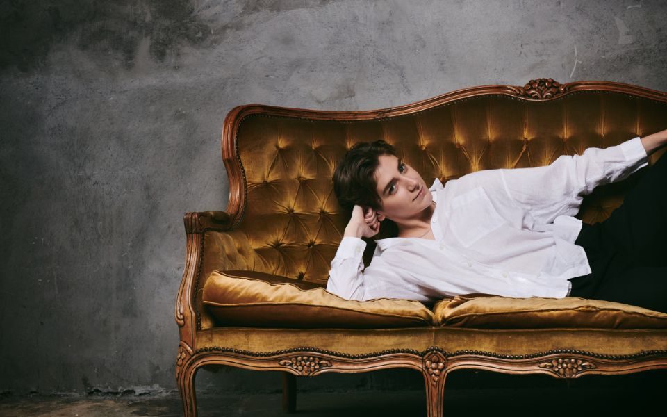 Sarah Keyworth wearing a white cotton shirt lying on a gold velvet sofa with a grey background.