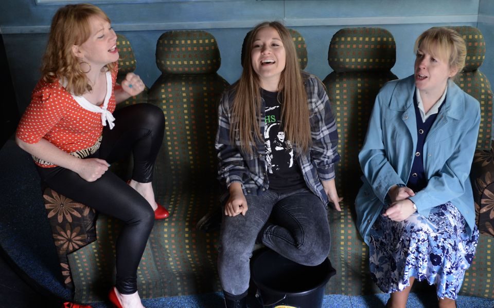 Three women sing together on bus seats.