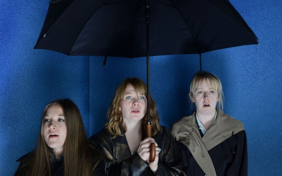 Three women look dramatically beyond the camera, under an umbrella.