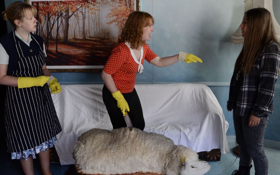 Three women stand around a sheep wearing rubber gloves. The central woman holds a knife.