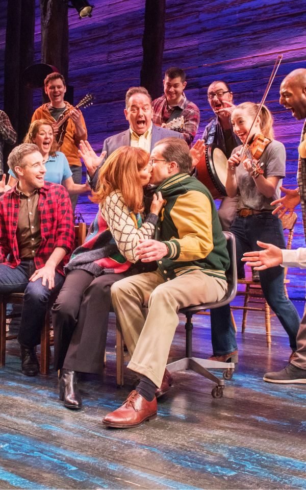 Two actors passionately kissing on stage. She's grabbing his green and yellow varsity jacket. They're surrounded by an excited-looking group of people and musicians with violins.