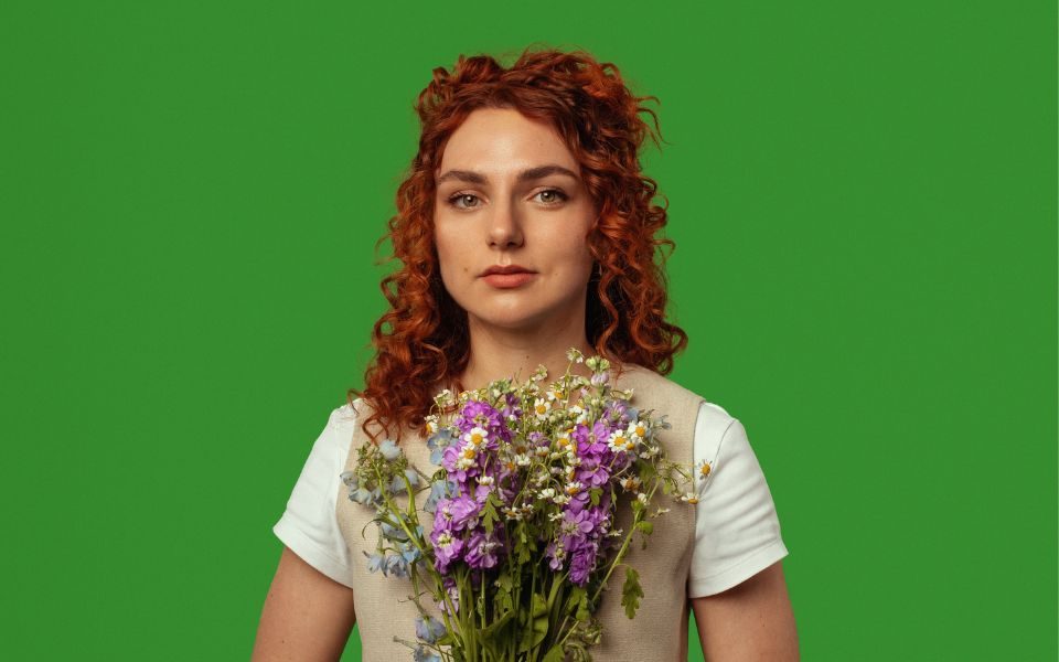 Ania Magliano in a beige waistcoat holding a bouquet of wild flowers to her chest against a green background.