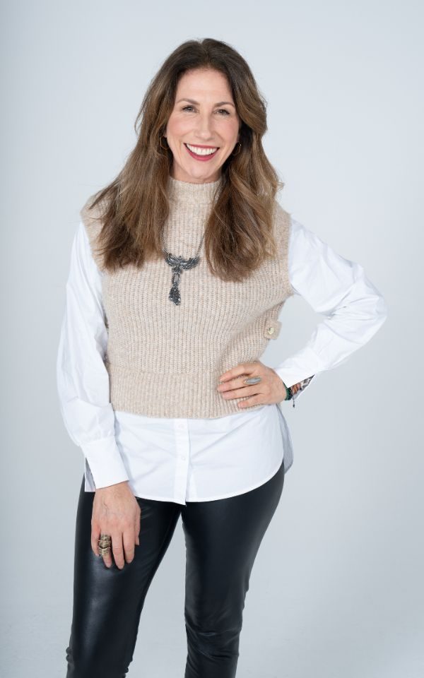 Gaynor Faye in a white shirt and beige jumper smiling at the camera with her hand on her hip