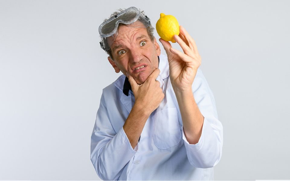 Henning Wehn in a white lab coat and safety goggles on his head holding a lemon in his left hand looking at it perplexed.
