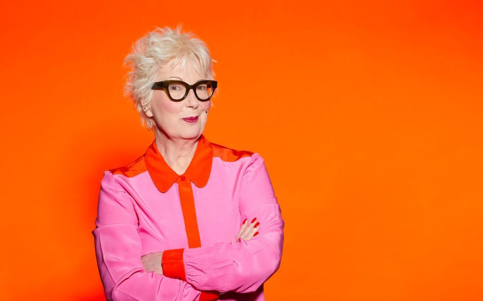 Jenny Eclair with her arms crossed wearing a pink and orange shirt and her signature black glasses against an orange background.