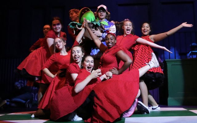 The youth theatre cast of Little Shop of Horrors wearing red dresses and posing for a photo around a boy holding up a plant puppet.