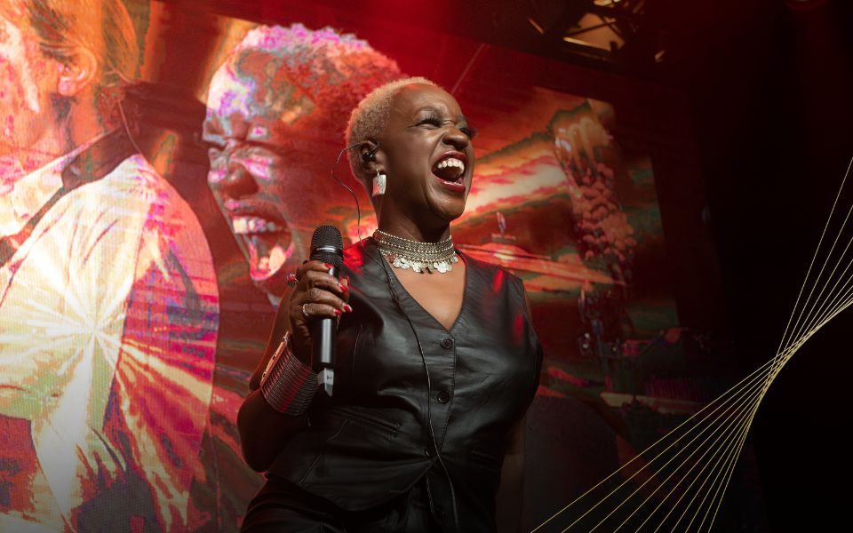 Woman looking very happy on stage wearing a black leather waistcoat and holding up a microphone in her right hand. She appears on the screen behind her.