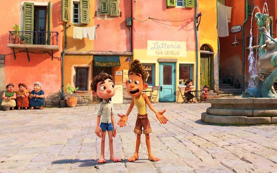 Still from Disney's Luca of Luca and Alberto stood on a cobbled street in Italy. Alberto has his hands out to the side in exclamation and brightly coloured terraced buildings are behind them and people sat at café tables and on benches, and a fountain is to the right of them