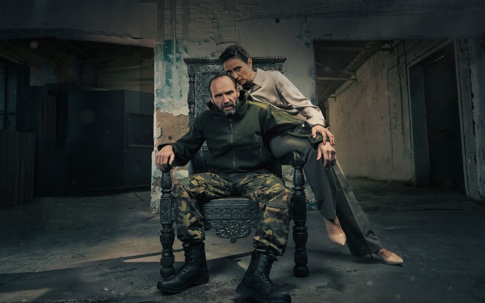 Ralph Fiennes as Macbeth sat on a metal throne wearing army camouflage trousers, dark green jacket and combat boots and Indira Varma as Lady Macbeth in a pale pink silk blouse, grey trousers and pale pink heels sat on the left arm of the chair. They are in an abandoned warehouse.