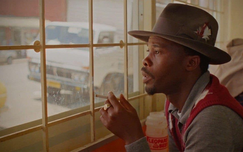 A still from Mapantsula of a man with a wide-rimmed hat, sweater vest and grey shirt smoking at a window.