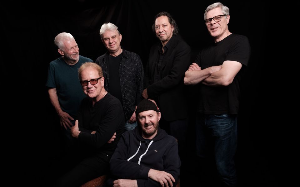 The six members of Oysterband wearing black on the top against a plain black background.