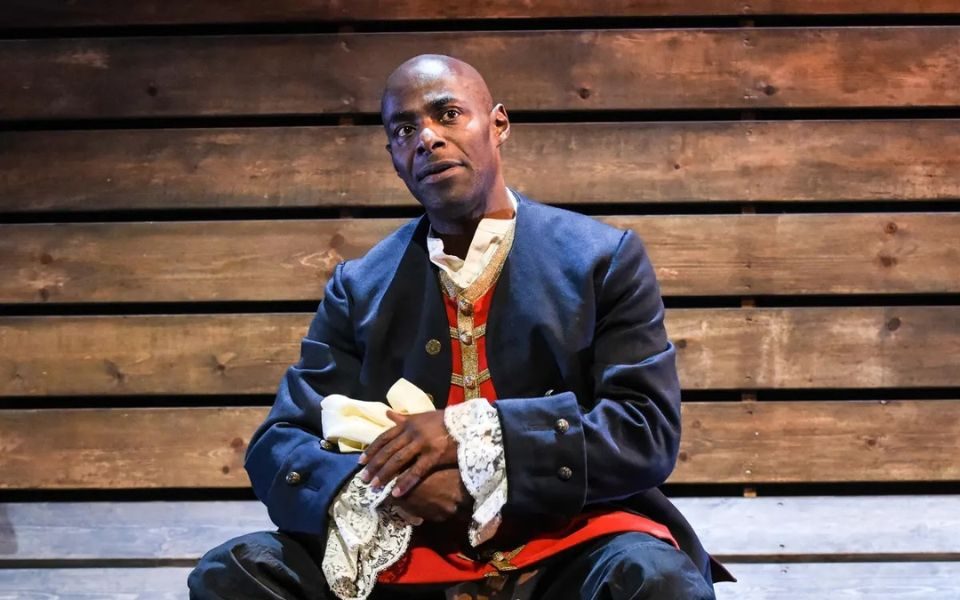Paterson Joseph dressed in blue and red 18th Century military uniform sat against some wooden plans.