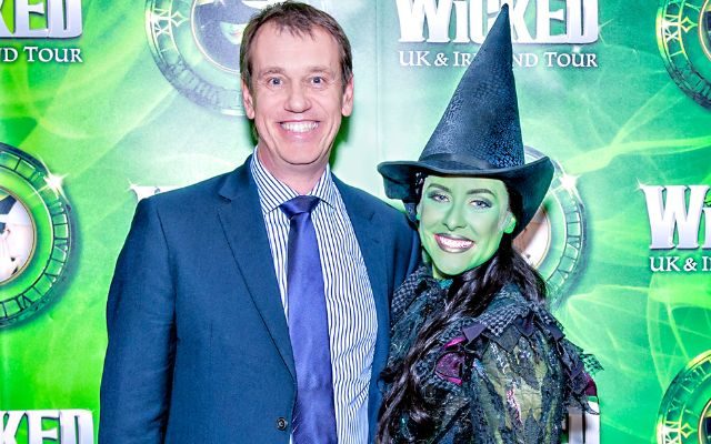 Chris Blythe in a suit smiling at the camera with Amy Webb dressed in a black witch outfit as Elphaba from Wicked in front of the Wicked press board.