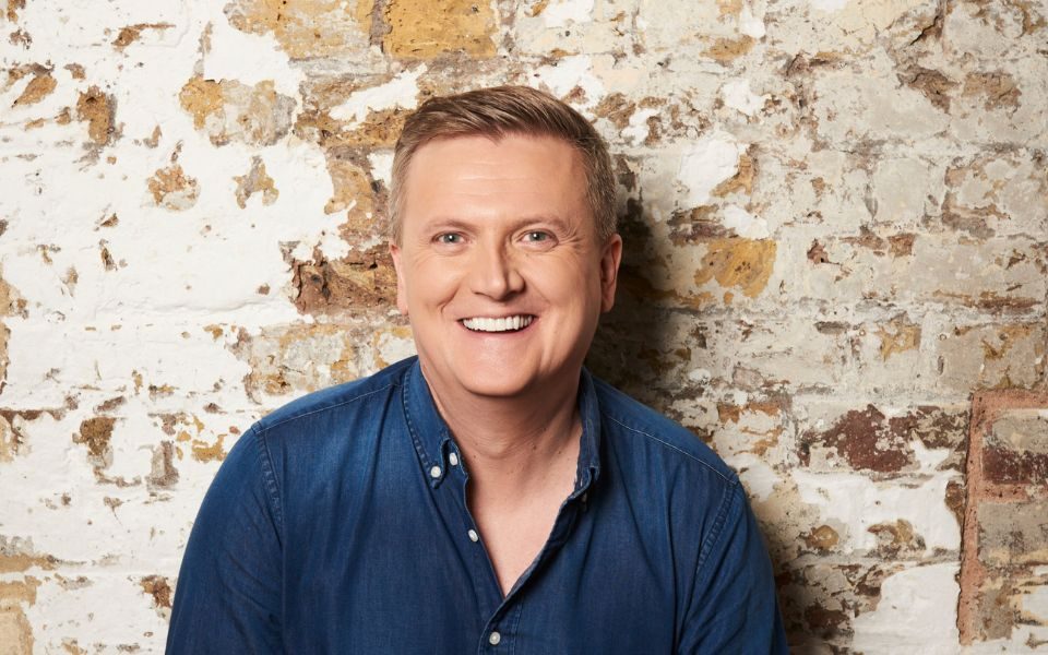 Aled Jones stood against an exposed brick wall smiling.