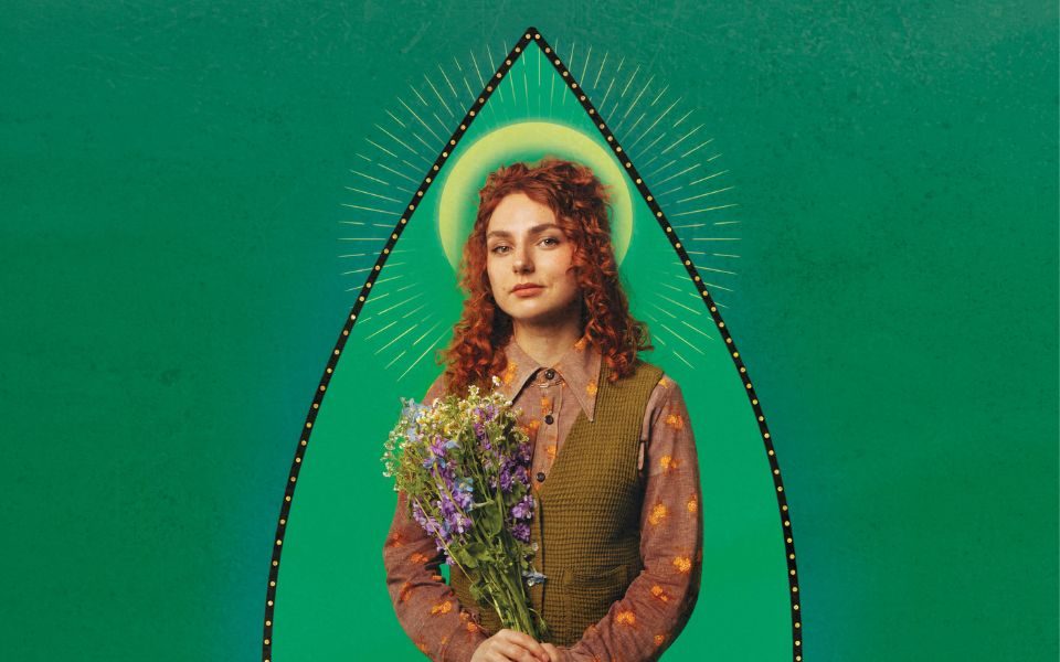 Ania Magliano with a church-painting-style halo in a green sweater vest and patterned shirt holding a bouquet of wild flowers to her chest stood in a the top of a tear-drop shape black outline against a two-toned green background.