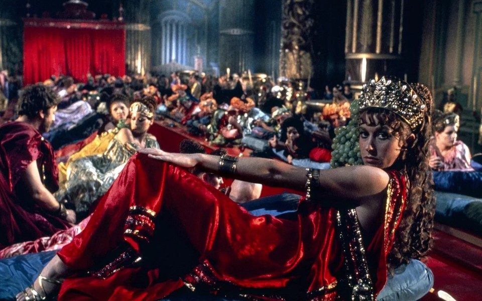 A still from Caligula of a room with a banquet table laid out with lots of people around it and on the the table in the blurry background and a woman in a red silk toga and golden crown lying in the foreground.
