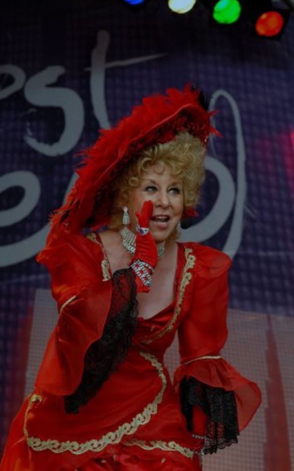 Jan Hunt performing in a red dress, hat and gloves, holding her hand up to her mouth as if whispering.