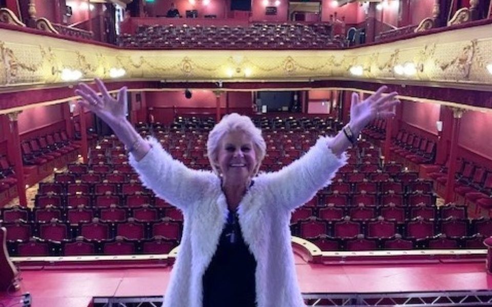 Jan Hunt with her arms out-stretched standing on the City Varieties stage with the auditorium in the background