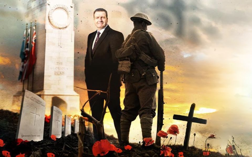 John Nichol in a suit stood next to a World War I soldier facing away from him, a cenotaph, war graves and crosses as you would see on top of a grave. Poppies appear at their feet.