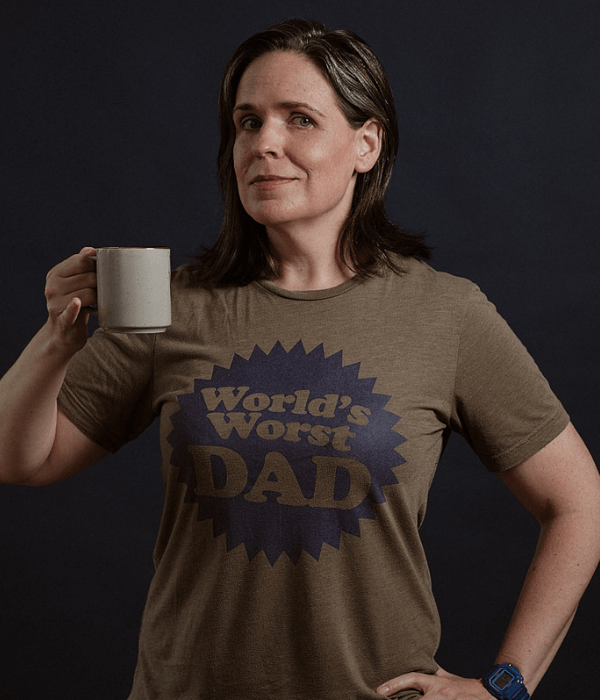 Kate McCabe looks towards the camera holding a mug and wearing a t-shirt saying 'World's Worst Dad'