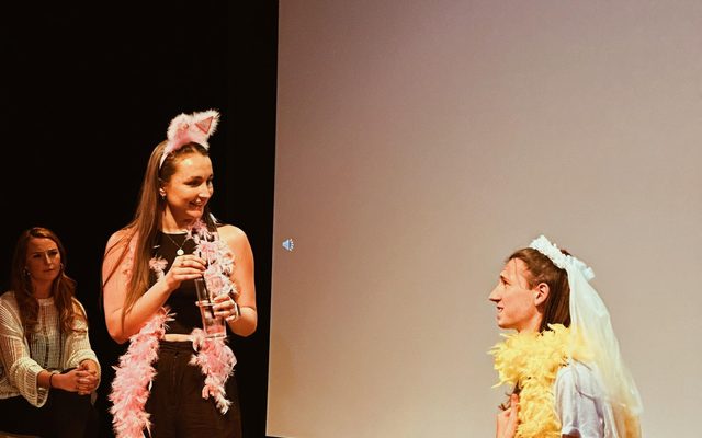 LAIT Method and Material Showcase One actor sits on a block in a feather boa and bridal headpiece, while another looks down at him also wearing a feather boa