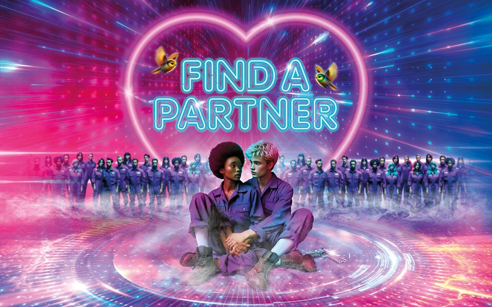 A couple sat in a space-age circular platform wearing purple boiler suits holding each other and looking at each other. A sparkly silver gun is next to them. Behind them are two rows of other people wearing the same boiler suits and a screen reading Find A Partner in blue neon text surrounded by two hummingbirds and a pink neon heart.